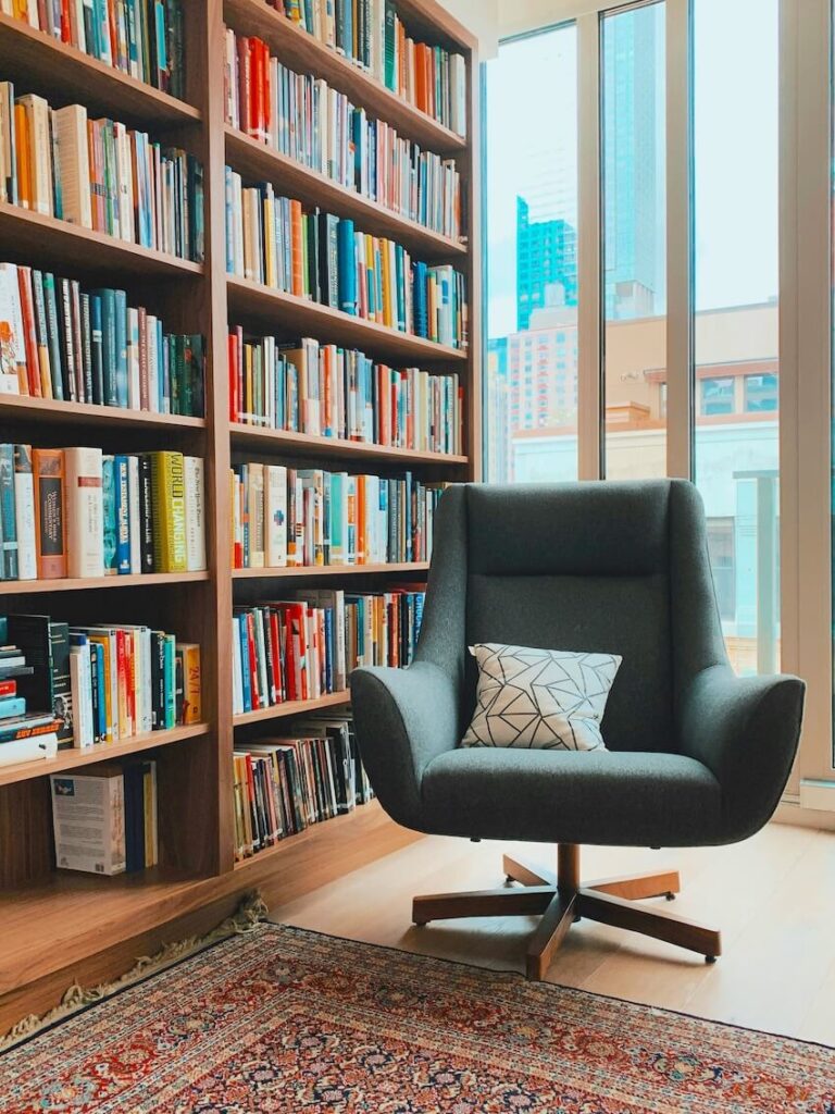 A cozy armchair situated in front of a floor-to-ceiling bookshelf filled with English resources and a large window overlooking a city skyline.